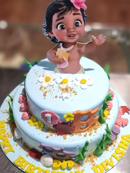 An adventurous Moana-themed cake for a first birthday. This two-tier cake is decorated with tropical flowers, seashells, and a baby Moana topper.