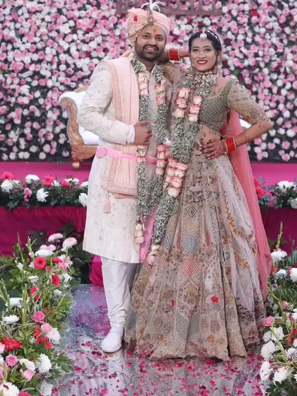 The happy couple posing against a beautiful floral backdrop. Her makeup looks fresh and radiant, perfect for their daytime wedding ceremony.