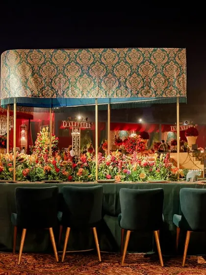 A long dining table with an overhead canopy featuring intricate patterns. The deep teal velvet chairs and lush florals create a luxurious and inviting space for guests to dine.