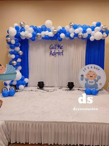 A stage setup for a Baptism or naming ceremony. This blue and white theme includes a balloon arch, a personalized sign, and cute baby character cutouts.