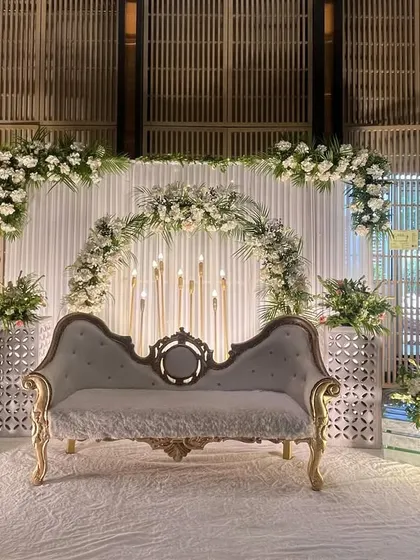 A sophisticated and elegant stage design. A classic grey vintage sofa is set against a backdrop of white drapes and white floral arches, accented with tall candle stands for a warm glow.