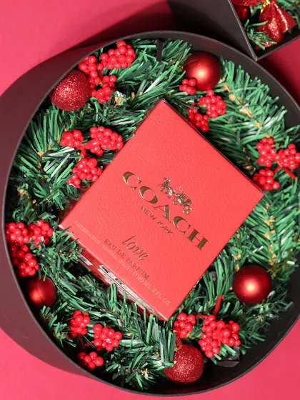 A luxury Christmas gift featuring a Coach "Love" perfume, nestled in a festive wreath of pine and red berries inside a sleek black box.