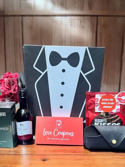 A dapper gift set for him, presented in a fun tuxedo-themed box. It includes a bottle of wine, Beardo perfume, "Love Coupons," Hershey's Kisses, and a stylish sunglasses case.