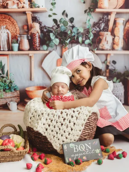 A creative "Mini MasterChef" session, with a big sister proudly hugging her newborn sibling, who is nestled in a basket.