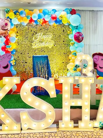 A Peppa Pig themed birthday party for a 4-year-old. The stage features a gold shimmer wall, character cutouts, and large marquee letters spelling "AKSHAJ".