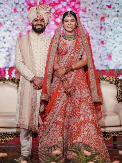 A classic full-length portrait of the bride and groom on their wedding day. Their happy smiles and elegant posture in front of a floral backdrop make for a perfect formal photograph.