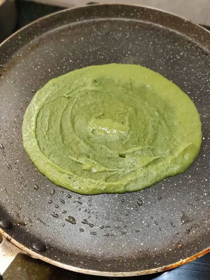 A spinach chilla being cooked on the pan, showing the fresh, green batter spread out to form a thin crepe.
