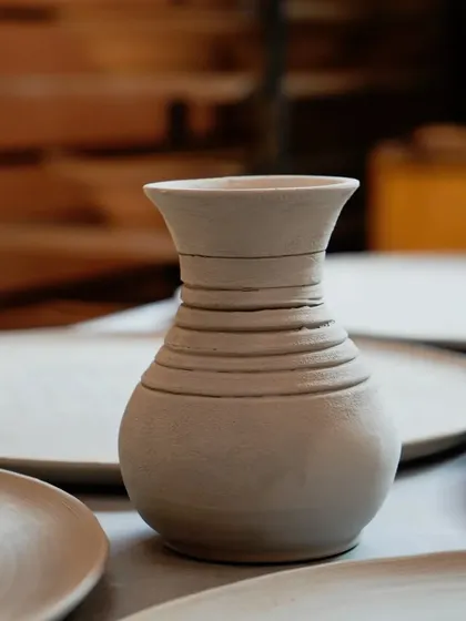 A single, unfired vase with carved lines, sitting amongst flat plates in our studio. This image captures a quiet moment in the creation process.