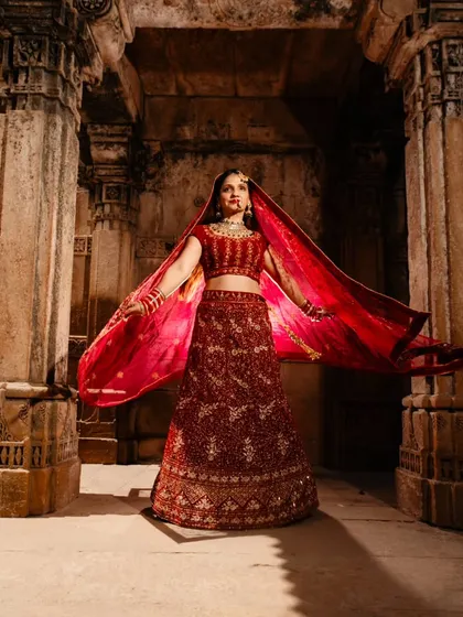 A full-length portrait of the bride, showcasing the magnificent detail of her red lehenga against the ancient stone. The flowing dupatta adds a touch of grace and movement to this regal shot.