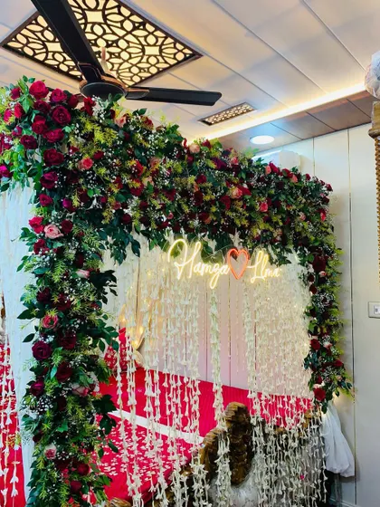 A lush and green welcome bride setup. The bed is framed by a dense arch of greenery and red roses, with a custom neon sign and hanging floral strings.