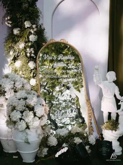 An elegant welcome sign for an engagement ceremony, etched onto a gold-framed mirror and surrounded by lush white flowers and classical statues.
