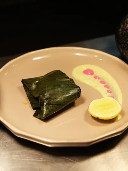 A dish of fish steamed in a banana leaf, presented with a colourful sauce and a slice of lemon. The plating is clean and elegant, allowing the natural beauty of the ingredients to shine.