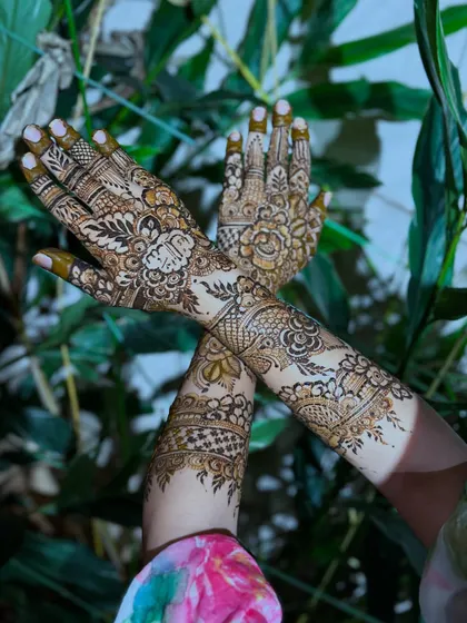 A crossed-hands pose set against a natural green background, making the rich brown of the henna stain stand out beautifully.