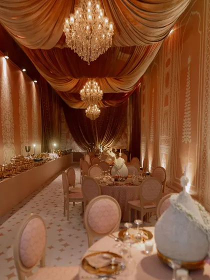 A view of the dining hall, where the ceiling is draped in soft, flowing fabrics and lit by crystal chandeliers. The walls are adorned with custom panels featuring traditional Indian motifs.