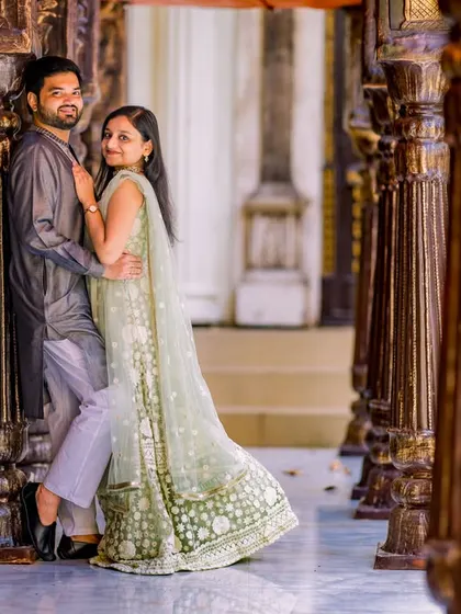 A stylish pose in a corridor of wooden pillars. The bride's flowing green lehenga and the groom's grey kurta create an elegant and modern traditional look.