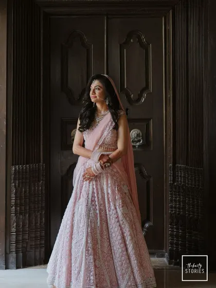 An elegant full-length portrait of the bride in a stunning pink lehenga, standing before a grand wooden door.