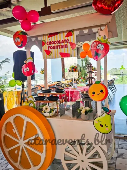I incorporate engaging food experiences like this chocolate fountain cart, decorated with smiling fruit balloons and a custom sign, making snack time an activity in itself.