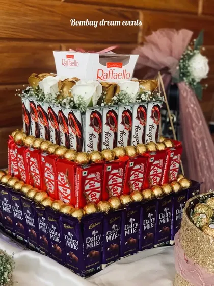 A multi-tiered chocolate tower made with Dairy Milk, KitKat, Ferrero Rocher, and Raffaello, topped with white roses. This is a stunning and delicious centerpiece for any celebration.