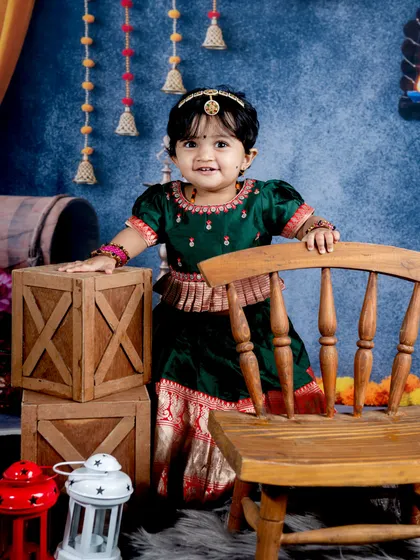 A beautiful toddler dressed in a traditional green and red South Indian outfit. The festive backdrop with hanging flowers and diyas is perfect for celebrating cultural occasions.