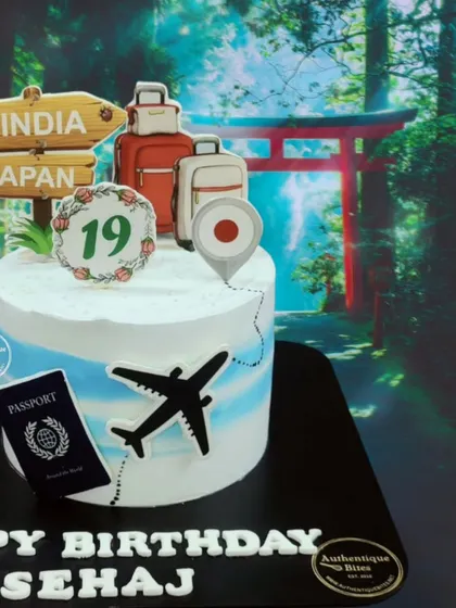 A travel-themed cake for someone heading to Japan. The design includes a passport, an airplane, luggage, and a signpost pointing from India to Japan.