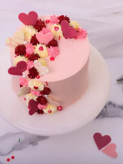 We craft a cascade of love with this romantic anniversary cake. A beautiful arrangement of red, pink, and white chocolate hearts and buttercream flowers flows down the side of a soft pink cake, creating a design that is bursting with affection.