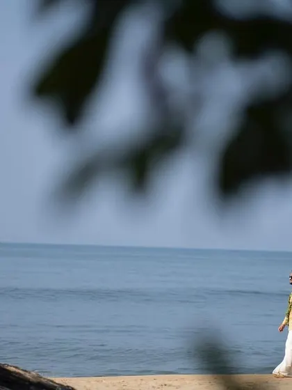 A wide shot of a couple walking hand-in-hand along a sandy beach, with the vast ocean beside them. The use of foreground blur adds an artistic, observational feel to this pre-wedding photograph.