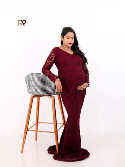 A confident solo portrait. The mom-to-be poses with a stool, her deep red lace gown creating a look of modern sophistication against the clean white background.