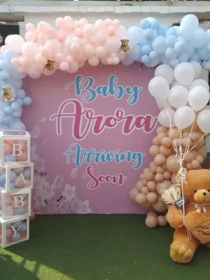 An outdoor gender reveal setup with a "Baby Arora Arriving Soon" theme. I created a large balloon arch in pastel pink and blue around a custom backdrop, adding 'BABY' boxes and a large teddy bear prop.