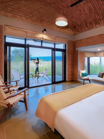 This is a bedroom in the Waghoba Ecolodge, a space designed for tranquility and immersion in nature. The vaulted ceiling, made from conical pottery tiles, and walls of stabilized adobe blocks create a cool, earthy sanctuary, while the large window opens directly to the restored forest landscape of Tadoba.