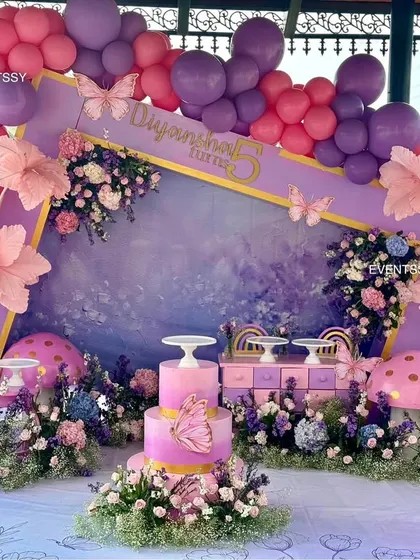 A full view of the purple and pink floral party, showing the cake table and dessert stands integrated into the enchanting scene. The hand-painted backdrop adds an artistic, dreamy quality.