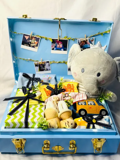 A gift hamper for a baby boy, packed in a beautiful blue trunk. It includes a soft elephant toy, a string of photos, and neatly wrapped clothing essentials.