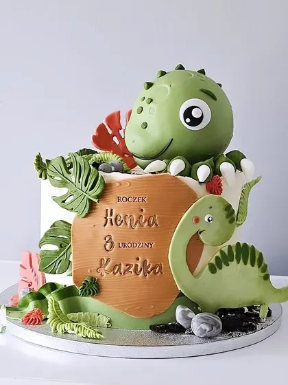 A fun and friendly dinosaur-themed cake for a child's birthday. This design features a large, cute green dinosaur topper and a smaller dino friend, surrounded by tropical leaves.