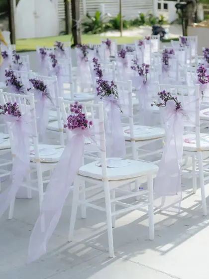 White chairs decorated with delicate purple flowers and ribbons, ready for guests at a pastel-themed wedding ceremony.