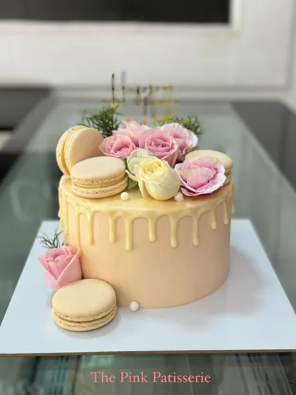 A lovely peach-colored drip cake topped with a beautiful arrangement of fresh pink and yellow roses, macarons, and delicate greenery.