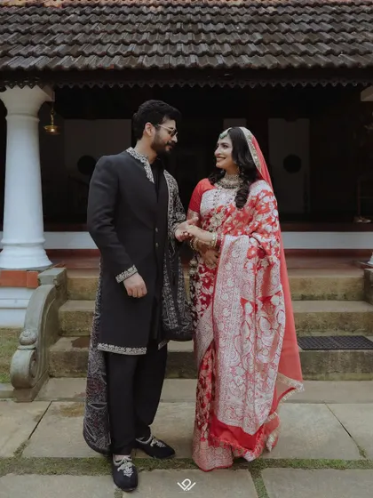 A couple in beautiful red and black traditional outfits, looking at each other lovingly in front of a heritage building.