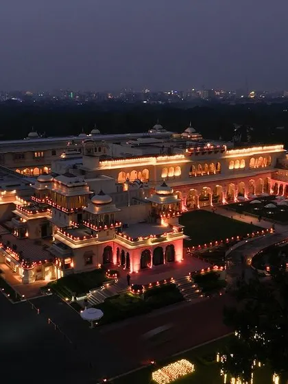 The Rambagh Palace in Jaipur, beautifully illuminated at dusk for a royal wedding event. This aerial shot captures the sprawling heritage property and the warm, inviting lights of the celebration, setting a luxurious and elegant tone.