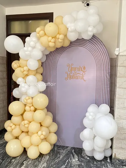 An elegant 'Umrah Mubarak' decoration to welcome loved ones. The setup features a lavender arched backdrop with a beautiful balloon garland in pastel yellow and white.