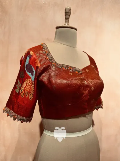 A red zari kota blouse with an intricate peacock design and floral embroidery on the neckline and sleeves.