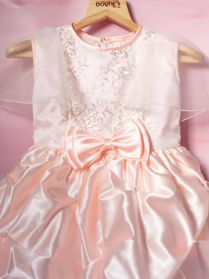A gorgeous party dress in a soft, peachy-pink satin. This design features a dramatic bubble hem, a large bow, and delicate floral embroidery on the bodice.