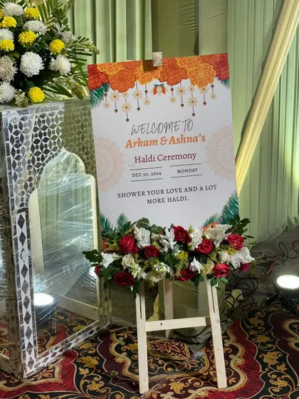 A personalized welcome sign for 'Arkam & Ashna's Haldi Ceremony', placed on an easel and decorated with fresh flowers.
