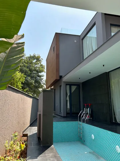 A narrow pool runs along the side of Gulmohar Farmhouse, making efficient use of the space while providing a refreshing visual element. The modern facade with its mix of wood and grey texture provides a sophisticated backdrop.