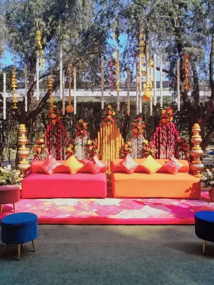 A vibrant Mehendi seating area with bright orange and pink sofas, set against a backdrop of colorful floral strings.