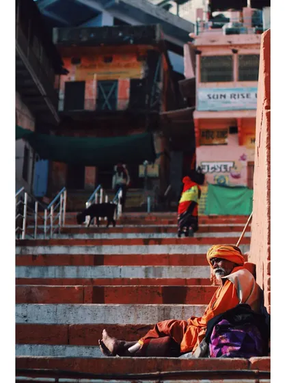A sadhu rests on the steps of a ghat, bathed in the warm morning light. The vertical composition emphasizes the layers of life happening on the ghats, from the resting man to the people in the background.