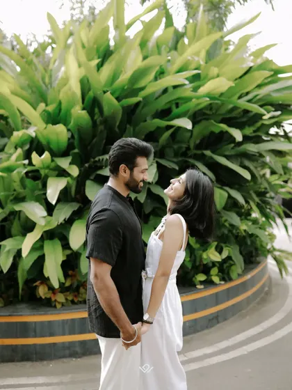 A happy, candid moment between a couple in casual attire, holding hands against a backdrop of green foliage.