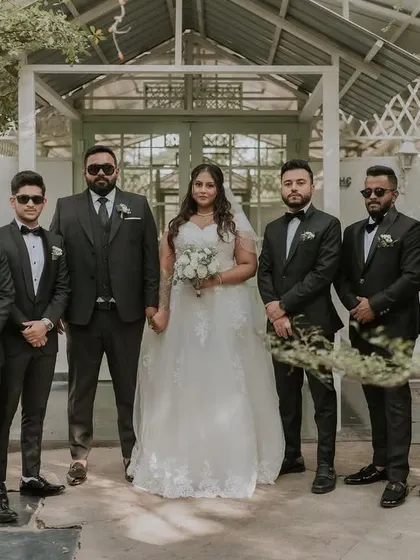 A bride and her groomsmen pose for a classic group photo. The clean, white architecture of the cottage provides a great contrasting background for their dark suits.