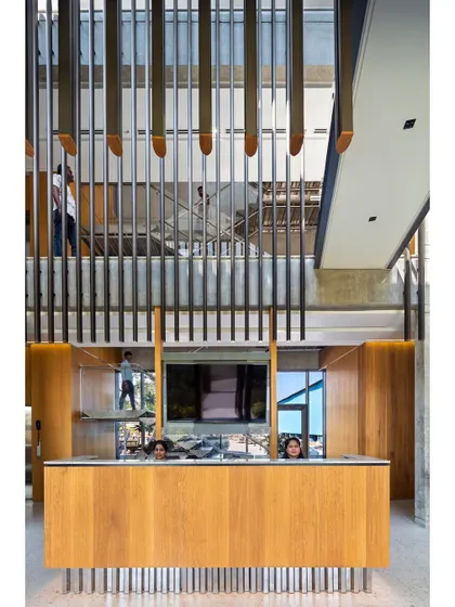 A frontal view of the reception desk, framed by the multi-story interior. The design uses ribbed oak paneling and exposed concrete to create a minimalist yet warm aesthetic.