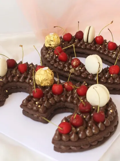 A creative number "3" shaped cake. This chocolate cake is piped with chocolate buttercream and decorated with fresh cherries, Ferrero Rocher, and macarons.