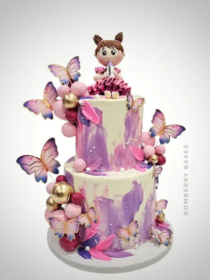 A two-tier butterfly-themed cake with a beautiful purple and pink watercolor effect, adorned with a cute girl figurine.