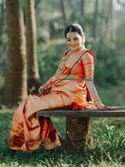 A serene moment captured outdoors. The rich red of the Kanchipuram silk saree contrasts beautifully with the tranquil nature setting.