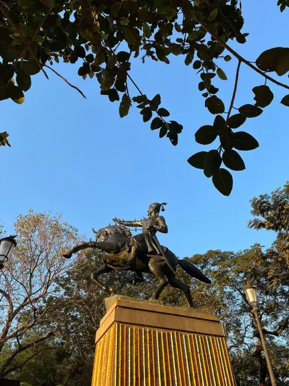 The majestic equestrian statue of Chhatrapati Shivaji Maharaj framed by tree leaves, with its base decorated with marigold garlands.